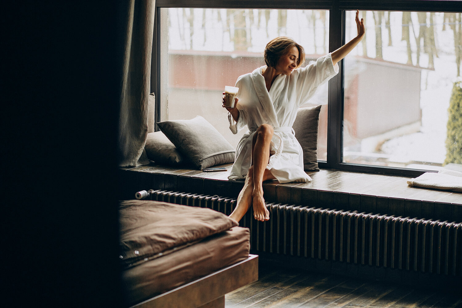 Femme en peignoir se relaxant avec une boisson chaude près d'une fenêtre après un soin spa ou un massage